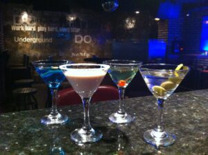Four cocktails lined up on a bar counter in front of a neon-lit 'Underground' wall mural