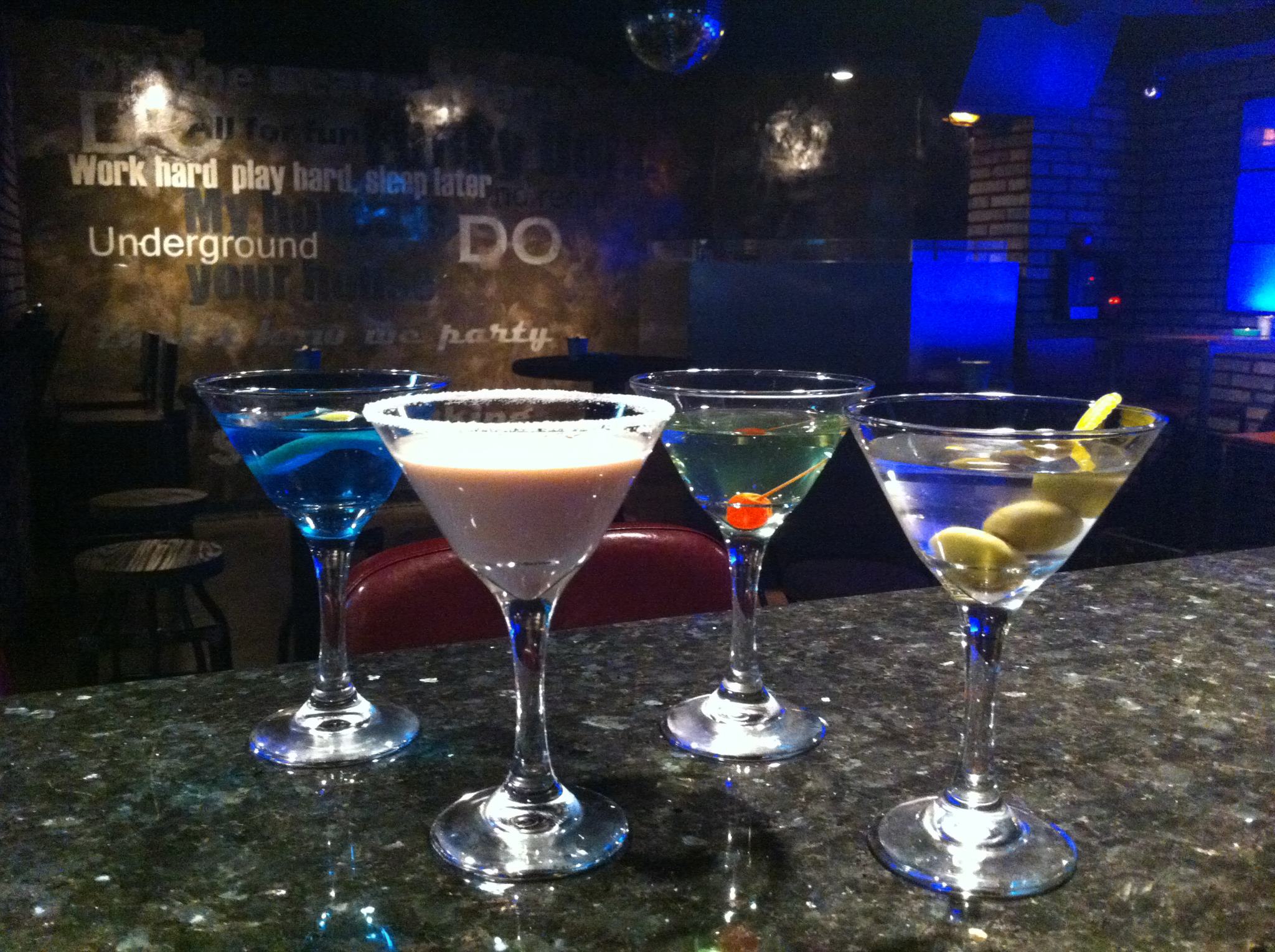 Four cocktails lined up on a bar counter in front of a neon-lit 'Underground' wall mural