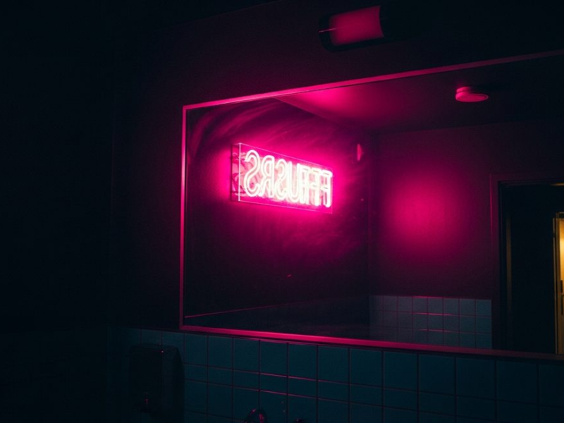 Dark club bathroom mirror reflecting hot pink neon signage, moody late-night atmosphere with gold and teal accents