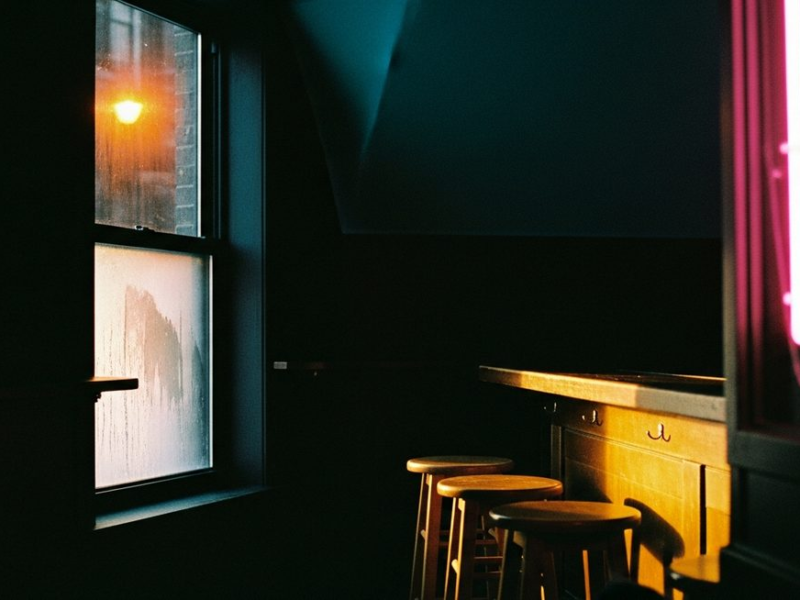 Small warm club interior in winter, condensation on dark window with cold streetlight outside, golden bar light on worn stools, pink neon out of focus
