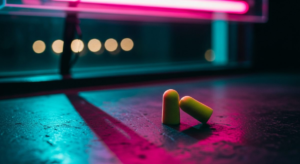 Foam earplugs resting on a dark surface, hot pink neon light casting a long shadow, blurred club lights out of focus in background