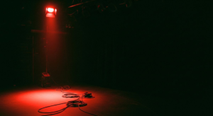 Empty stage under a single harsh red work light, cables coiled on the floor, deep darkness consuming most of the frame