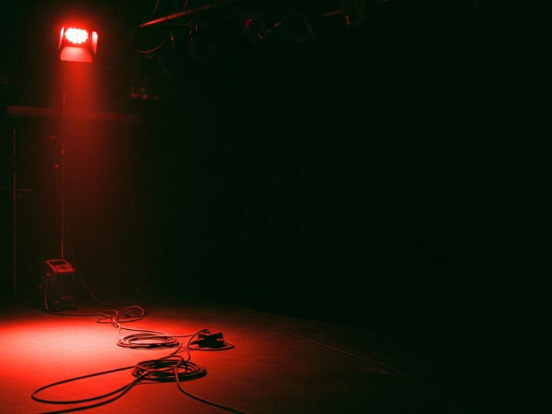 Empty stage under a single harsh red work light, cables coiled on the floor, deep darkness consuming most of the frame