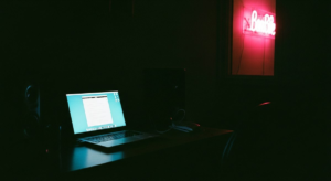 Laptop screen glowing in a completely dark room, hot pink neon reflection from outside window, teal light on ceiling above