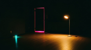Small dark practice room with warm gold standing lamp, hot pink light bleeding under a closed door, teal power strip glow on floor left