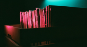 Vinyl record sleeves in a wooden crate, hot pink light raking across spines creating long shadows, teal glow from behind