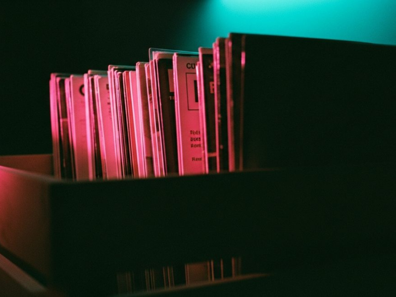 Vinyl record sleeves in a wooden crate, hot pink light raking across spines creating long shadows, teal glow from behind