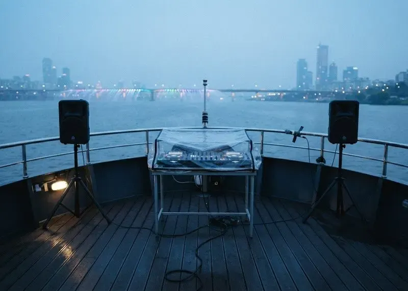 A small DJ rig under a weather cover at the stern of a Han River cruise boat at blue hour, two speakers on stands, the Seoul skyline across the water