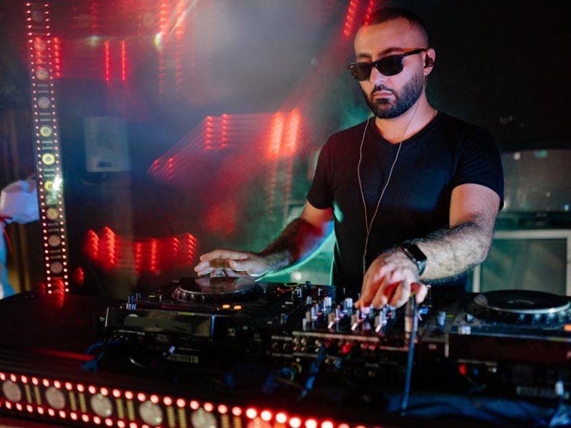A DJ wearing sunglasses adjusting a mixer between two CDJs under red lighting