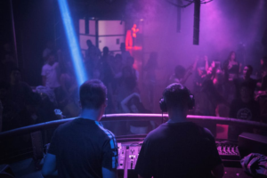 Two DJs viewed from behind performing at a mixer booth overlooking a dancing crowd
