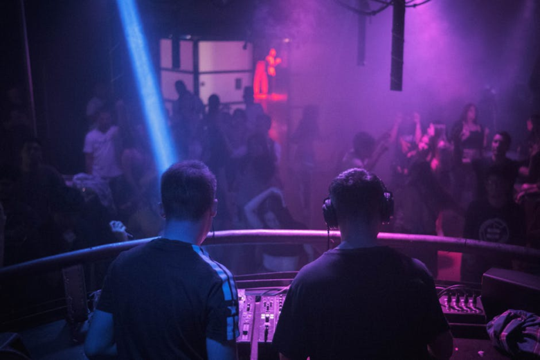 Two DJs viewed from behind performing at a mixer booth overlooking a dancing crowd