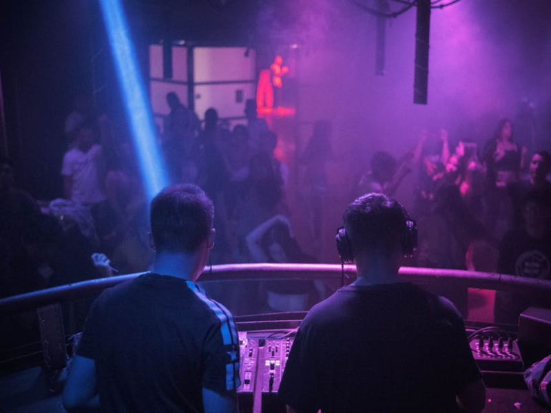 Two DJs viewed from behind performing at a mixer booth overlooking a dancing crowd