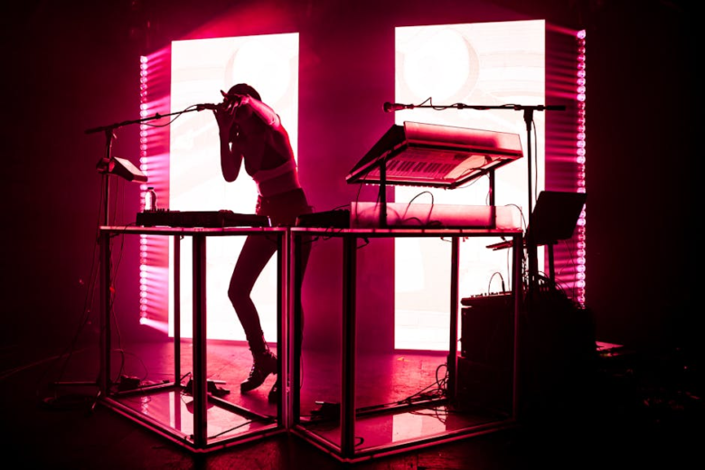 Silhouette of a performer singing into a microphone beside a keyboard under pink stage lights