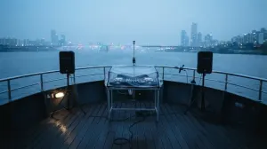A small DJ rig under a weather cover at the stern of a Han River cruise boat at blue hour, two speakers on stands, the Seoul skyline across the water