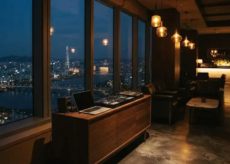A small DJ setup on a sideboard in a dim Seoul rooftop lounge, amber pendants, the Han River and city lights through the glass