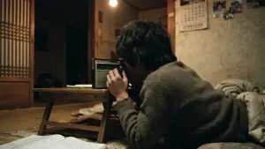 A young listener seen from behind holding a transistor radio to their ear in a 1970s Seoul home