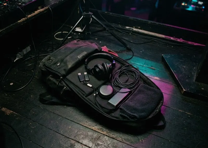 An open DJ backpack on a dark booth floor with headphones, USB sticks and earplugs