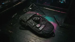 An open DJ backpack on a dark booth floor with headphones, USB sticks and earplugs