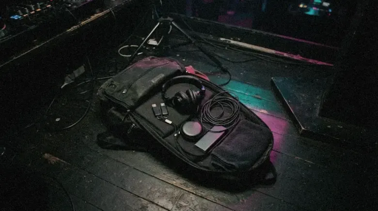 An open DJ backpack on a dark booth floor with headphones, USB sticks and earplugs