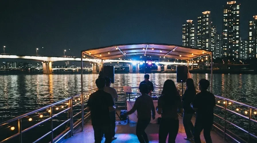 Guests dancing on the rear deck of a Han River boat at night, bridge lights behind