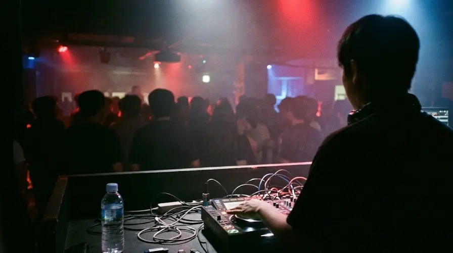 A DJ seen from behind at a small Seoul club booth, red and blue stage lights over a dark crowd