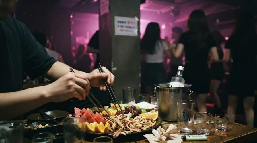 An anju platter on a Seoul lounge club table at 2am, people dancing in the aisle behind