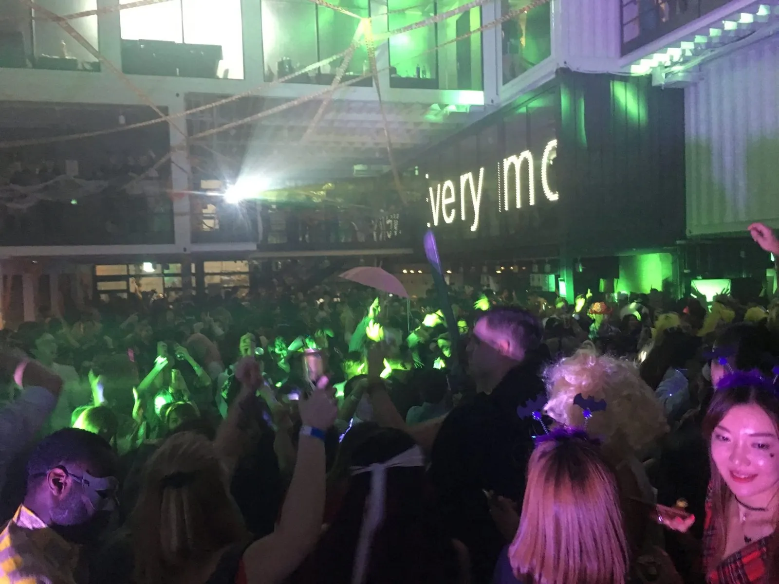 A costumed crowd dancing under green lights at a Halloween party in a shipping container venue