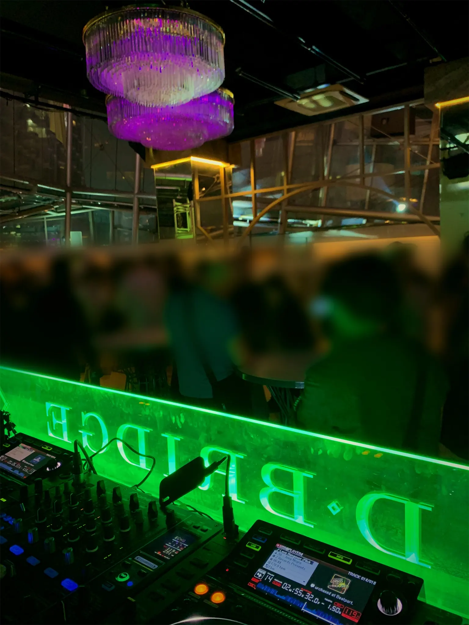 A DJ mixer behind a green lit glass panel, looking out over a Gangnam lounge crowd under chandeliers.
