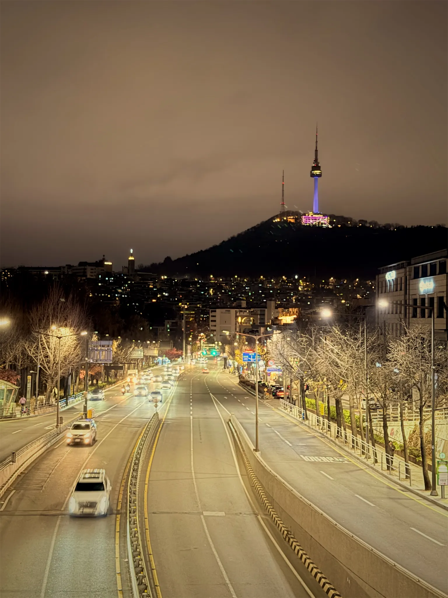 A wide road at night curving toward Namsan, with car lights, street lamps and N Seoul Tower lit on the hilltop.