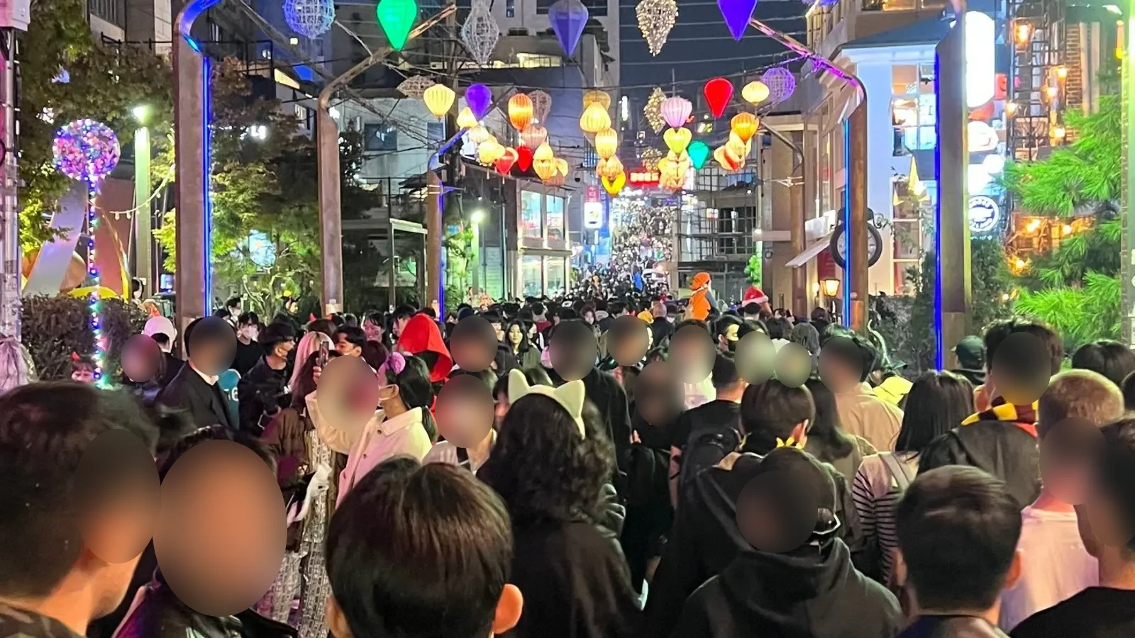 A packed Itaewon street on Halloween night under strings of coloured lanterns, people in cat ears and costumes, faces in front blurred