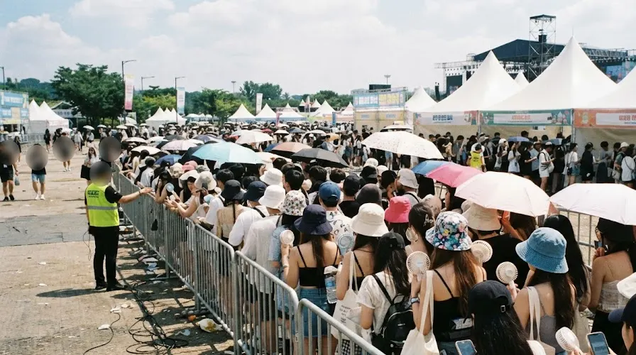 A queue of festival-goers with umbrellas and handheld fans at a Korean festival wristband exchange